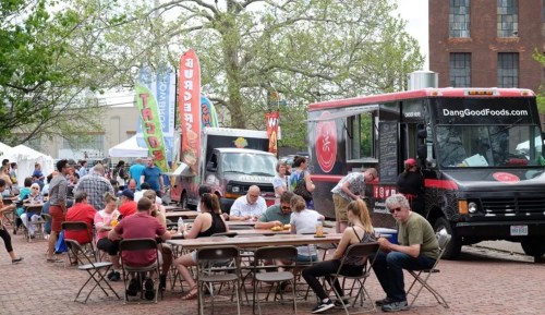 A bustling food truck festival with people enjoying meals at tables and colorful banners in the background.