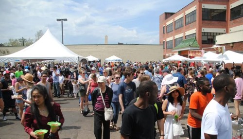A large crowd gathers at an outdoor event with tents, enjoying food and socializing on a sunny day.