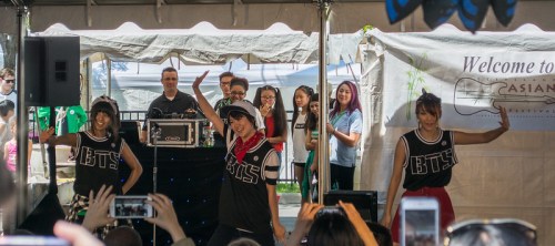 A group of dancers performs on stage at an outdoor event, with an audience capturing the moment on their phones.