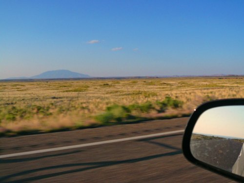 A view of a vast, open landscape with mountains in the distance, seen from a car's side mirror.