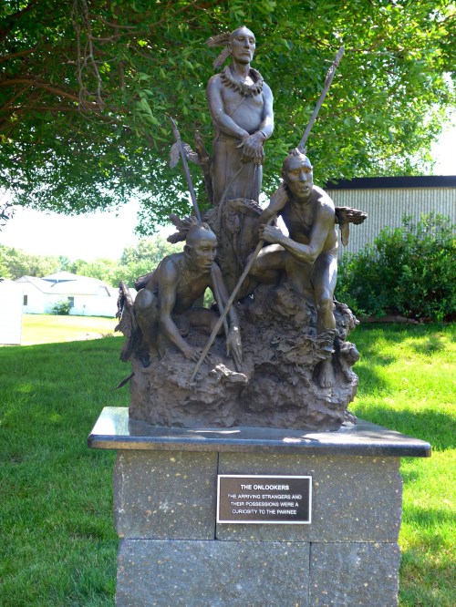 A bronze sculpture of three figures, two standing and one seated, surrounded by natural elements, with a plaque below.