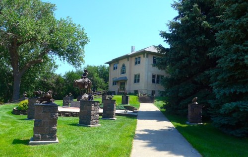 A large house surrounded by green grass and several bronze sculptures on stone pedestals under a clear blue sky.
