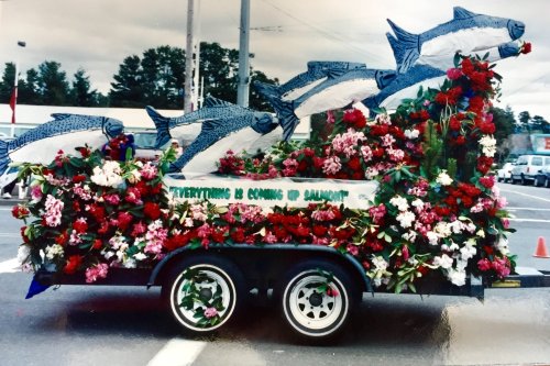 A colorful parade float decorated with flowers and large fish sculptures, featuring the phrase "Everything is Coming Up Salmon."