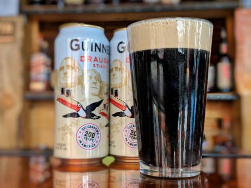 A glass of dark stout beer with a creamy head, next to two cans of Guinness Draught Stout on a wooden bar.