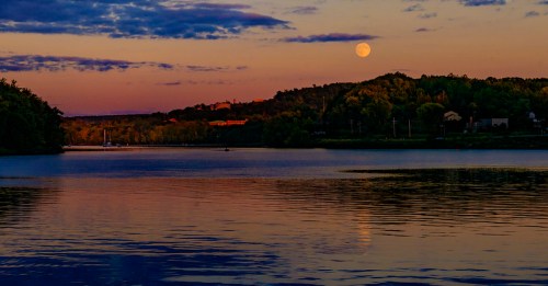 A serene river scene at sunset, with a full moon reflecting on the water and hills in the background.