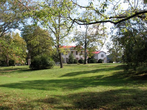 A large white house with a red roof surrounded by trees and a green lawn on a sunny day.