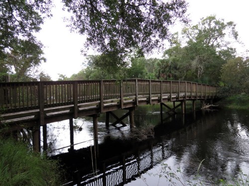A wooden bridge spans a calm river, surrounded by lush greenery and trees. Reflections shimmer on the water's surface.
