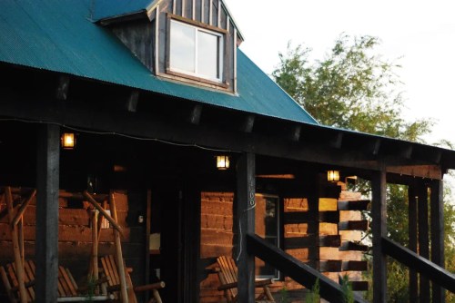 A rustic log cabin with a green roof, featuring a porch and large windows, surrounded by greenery.