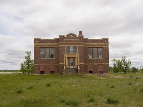 Abandoned brick school building with large windows, surrounded by grassy field under a cloudy sky.
