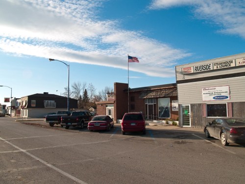 A small town street scene featuring a restaurant, parked cars, and an American flag against a clear blue sky.