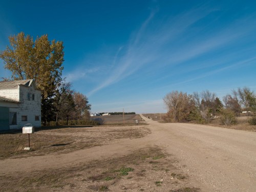 A dirt road leads into the distance, flanked by trees and an old house under a clear blue sky.