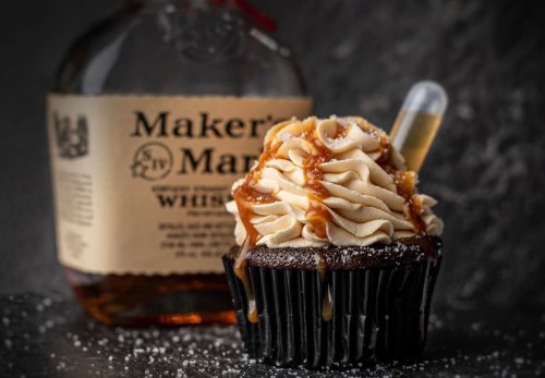 A chocolate cupcake topped with swirled frosting and caramel, next to a bottle of Maker's Mark whiskey.