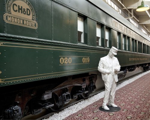 A vintage green train car with a white statue of a conductor standing beside it, set in a museum environment.