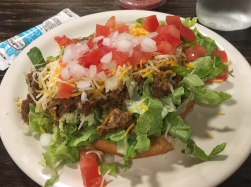 A plate of a taco salad topped with ground beef, lettuce, tomatoes, cheese, and guacamole on a crispy shell.