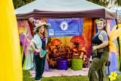 Two women in fairy costumes stand in front of a colorful booth with a whimsical backdrop at a festival.