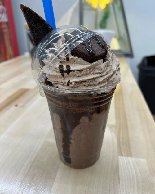 A chocolate milkshake topped with whipped cream, chocolate drizzle, and a brownie piece, served in a clear cup with a blue straw.