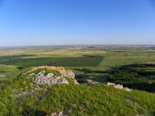 A panoramic view of rolling green hills and fields under a clear blue sky, seen from a rocky outcrop.