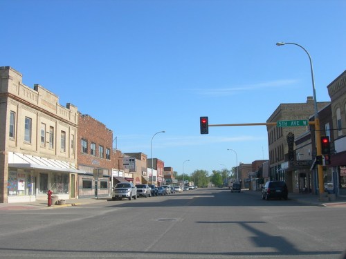 A quiet street view of a small town with brick buildings, parked cars, and a traffic light at the intersection.