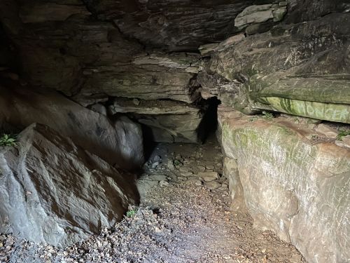 A dark cave entrance surrounded by large rocks and earthy terrain, with faint light illuminating the interior.