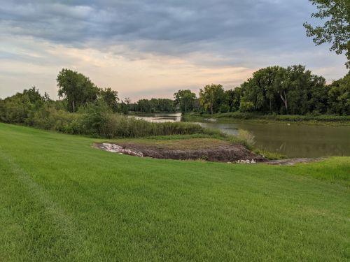 A serene landscape featuring a grassy area beside a calm river, with trees lining the shore under a cloudy sky.