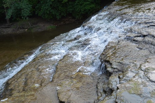 A gentle waterfall cascades over rocky terrain into a calm river, surrounded by lush green trees.