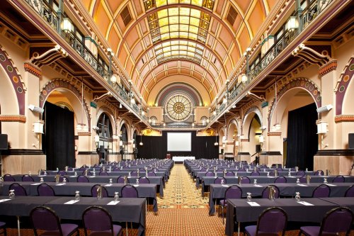 A spacious conference hall with rows of tables and chairs, featuring ornate architecture and a large screen at the front.