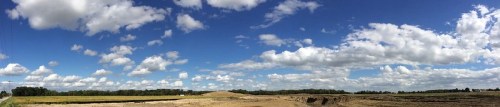 A panoramic view of a landscape with blue skies, fluffy clouds, and fields under construction.