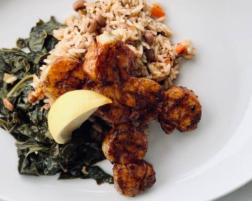 Grilled shrimp with a lemon wedge, served with rice and beans, and sautéed greens on a white plate.