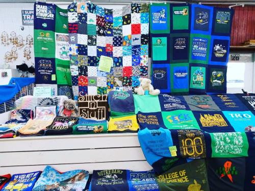 A colorful display of T-shirts and quilts, with a small dog resting on a table, showcasing various designs and patterns.