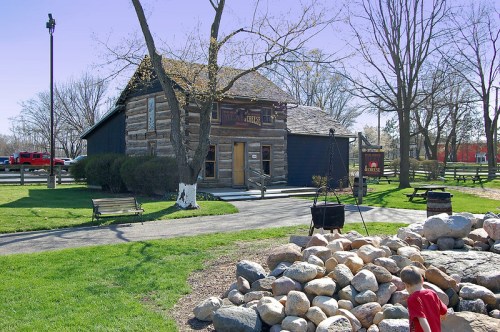 A log cabin-style building surrounded by green grass, trees, and a rock formation, with a child playing nearby.