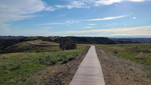 A winding pathway through grassy hills under a clear blue sky, with distant mountains visible on the horizon.