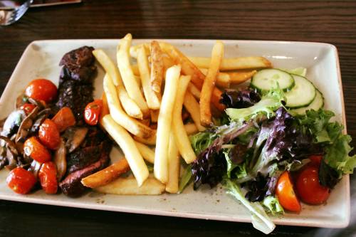 A plate with grilled steak, sautéed mushrooms and tomatoes, French fries, and a fresh salad with cucumbers and lettuce.