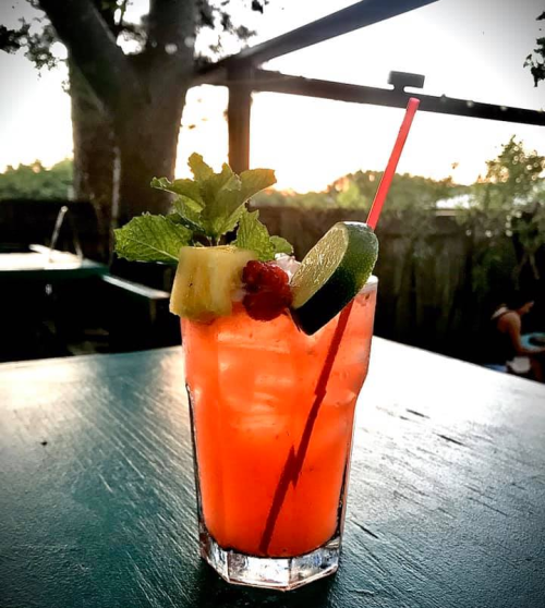 A vibrant cocktail garnished with mint, pineapple, lime, and raspberries, set against a sunset backdrop.