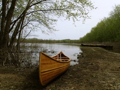 A wooden canoe rests on the shore of a calm river, surrounded by trees and greenery under a cloudy sky.