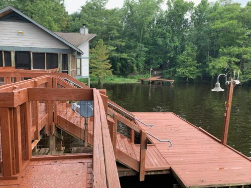 A wooden deck leads to a calm lake, surrounded by trees and a house in the background.