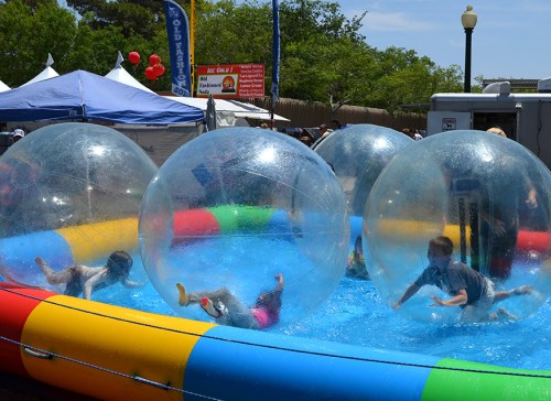 Children play inside large inflatable water balls in a colorful pool at an outdoor event.