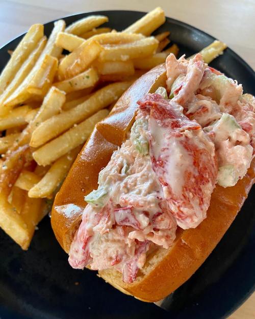 A lobster roll filled with creamy lobster salad, served with a side of golden French fries on a black plate.