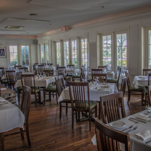 A spacious restaurant with wooden tables, white tablecloths, and large windows letting in natural light.