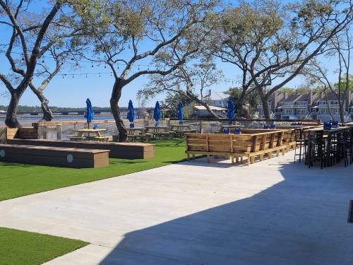 Outdoor seating area with picnic tables and umbrellas, surrounded by trees and a view of the water. Clear blue sky.