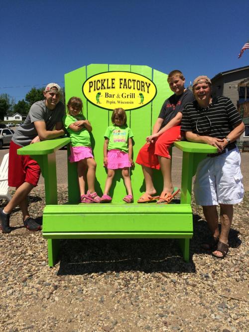 Five people pose together in front of a bright green "Pickle Factory" sign in Pippin, Wisconsin, on a sunny day.