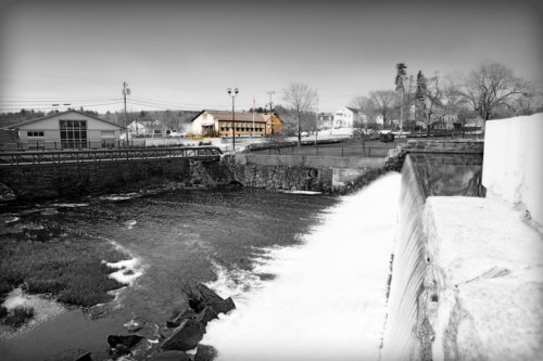 A black-and-white image of a river with a dam, featuring buildings and trees in the background.