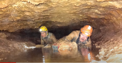 Two cavers in helmets navigate a narrow, water-filled cave passage, covered in mud.