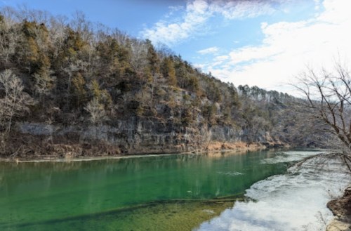 A serene river flows through a wooded landscape, with rocky cliffs and a clear blue sky above.