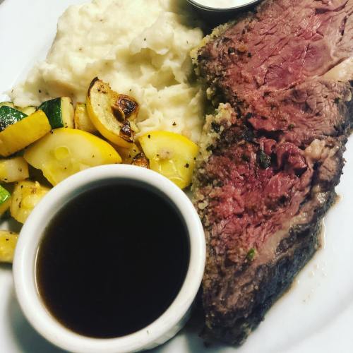 A plate featuring sliced beef, creamy mashed potatoes, sautéed zucchini, and a small bowl of sauce.