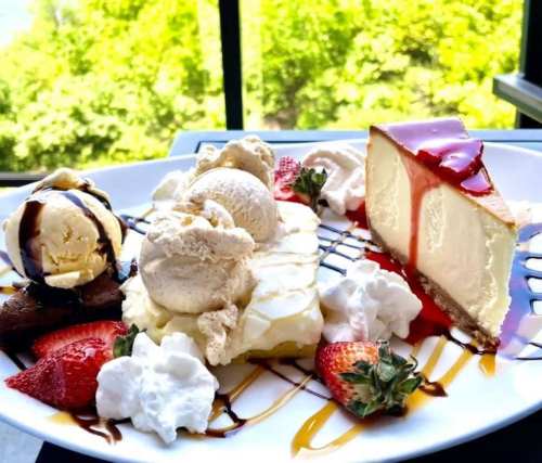 A dessert plate featuring cheesecake, ice cream, strawberries, whipped cream, and chocolate sauce, set against a green backdrop.