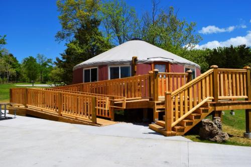A wooden ramp leads to a yurt surrounded by greenery under a clear blue sky.
