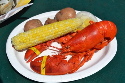 A plate with a cooked lobster, corn on the cob, and potatoes, served with a side of dipping sauce.