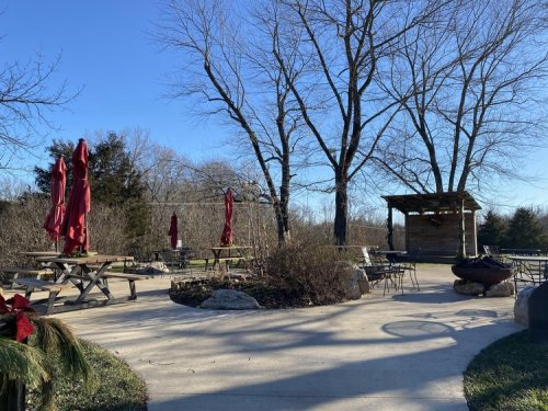 Outdoor seating area with picnic tables, red umbrellas, and a wooden gazebo surrounded by trees on a clear day.