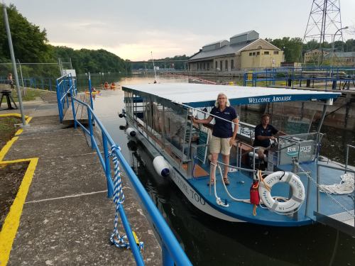 A tour boat docked at a lock, with two crew members on board and a scenic waterway in the background.