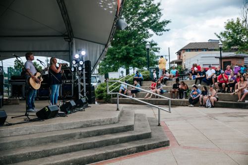 A musician performs on stage while an audience sits on steps, enjoying the outdoor event under a cloudy sky.